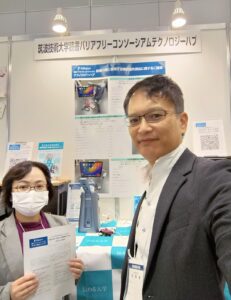 ブースの様子。機器を紹介するポスターや展示物の前で、本学スタッフが配布資料を手に映っている。